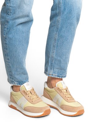 Light yellow and tan sneakers with suede and mesh panels, white accents, and brown rubber soles. Laces are beige, featuring a logo patch on the tongue.