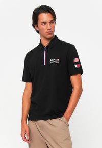Sort polo shirt med en kort lynlås, med "USA" og logoer på brystet og ærmet, der giver en blanding af bomuldsstruktur.