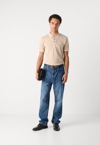 Beige polo skjorte med hvit krage, lyseblå jeans og svarte sko. Holder en svart veske, står foran en nøytral bakgrunn.