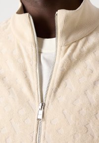 Beige texturerad zip-up jacka med ribbat krage över en vit t-shirt, visad på en person med mörk hudton.