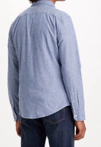 Jasno niebieska koszula z długim rękawem, wykonana z teksturowanej tkaniny, z kołnierzykiem zapinanym na guziki i mankietami, z zaokrąglonym dołem. Noszona z ciemnymi dżinsami.