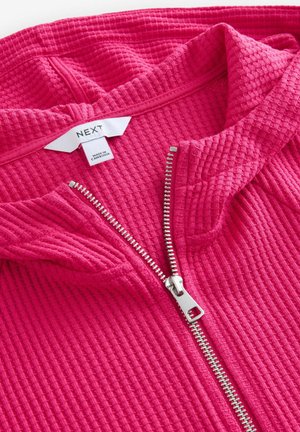 Fuksija zip-up dukserica izrađena od teksturiranog materijala. Sadrži srebrni zatvarač i oznaku na kojoj piše "NEXT" i "NASTANJENO U KAMBODŽI."