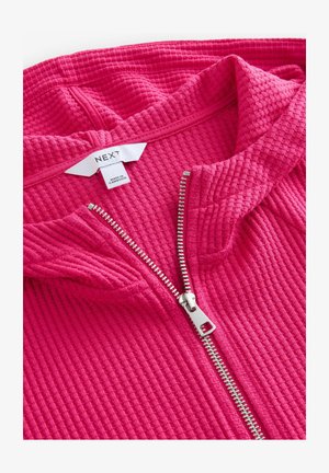 Fuchsiová zipová mikina vyrobená z texturované tkaniny. Obsahuje stříbrný zip a štítek s nápisem "NEXT" a "VYROBENO V KAMBODŽE."