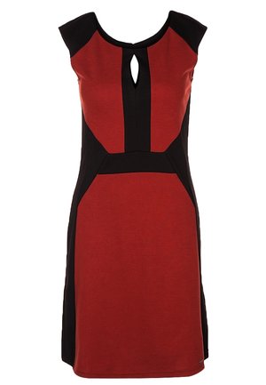 Vrouw draagt een rood-zwarte mouwloze jurk, zwarte enkellaarzen, doorschijnende zwarte panty, houdt een grote zwarte handtas vast.