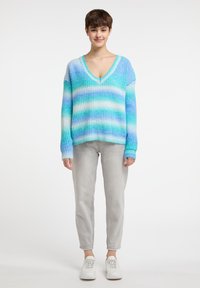 Maglione leggero e oversize con strisce gradientate blu e bianche, con un profondo scollo a V e texture a costine. Indossato con jeans grigi e sneakers bianche.