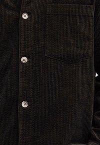 Tmavě zelená kordurová košile s vertikální jemnou texturou, vybavená náprsní kapsou a světlými knoflíky. Detailní záběr na látku a designové detaily.