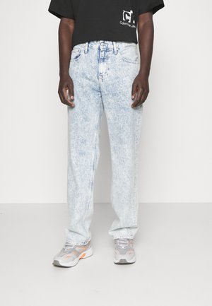 Jeans de mezclilla de lavado claro con un ajuste relajado, que presentan una textura desgastada, cinco bolsillos y un diseño de pierna recta, llevados con zapatillas grises.