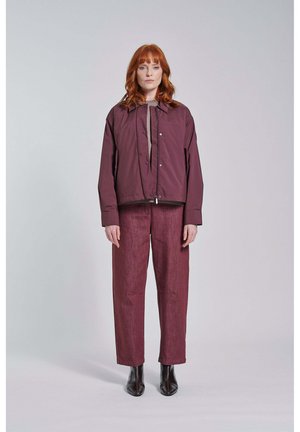 Vrouw met rood haar die een donkerpaarse jas, bijpassende losse broek en zwarte enkellaarsjes draagt, staand tegen een eenvoudige lichte achtergrond.
