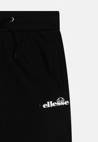 Crne pamučne trenirke s rebrastim strukom, zatvaranjem na vezicu i bijelim "ellesse" logom na lijevom bedru. Uključeni su bočni džepovi.