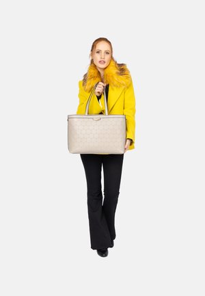 Beige tote taske med et tekstureret mønster og sort kant, holdt af en model iført en gul frakke med pelskrave, parret med sorte flared bukser.