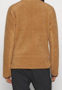 Brązowa polarowa kurtka o miękkiej, teksturowanej powierzchni. Posiada luźny krój i długie rękawy, z minimalnie widocznymi elementami metalowymi lub akcentami.