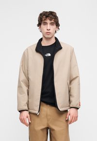 Μπεζ μπουφάν με μαύρο γιακά επενδεδυμένο με φλίς, κλείσιμο με φερμουάρ και ελαστικές μανσέτες. Φορεμένο πάνω από μαύρο T-shirt. Συνδυασμένο με χακί παντελόνι.