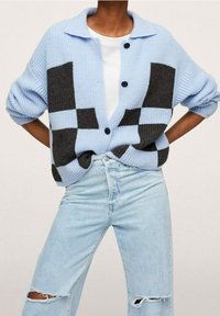 Cárdigan de punto azul claro con patrón de bloques gris oscuro, cierre de botones y textura suave. Combinado con jeans rasgados azul claro.