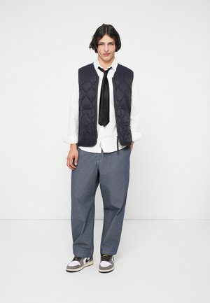 Marine blå quilted vest over en hvit skjorte med svart slips, kombinert med løse grå bukser og hvite sneakers med grå og svarte detaljer.