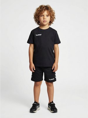 Junges Kind mit lockigem Haar, das schwarze Hummel-Sportkleidung und schwarze Sneakers trägt, steht auf einem schlichten Hintergrund und blickt nach vorne.