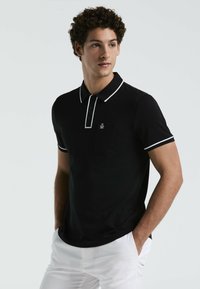 Schwarzes Poloshirt mit kontrastierendem weißen Kragen und Besatz, ausgestattet mit einer Vordertasche und einem Logo. Modell kombiniert mit weißen Hosen.