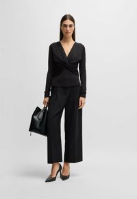 Maglietta nera a maniche lunghe con design frontale attorcigliato, abbinata a pantaloni neri a gamba larga. La modella tiene in mano una borsa nera. Scarpe nere con tacco alto.