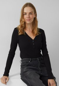 Czarna bluzka z długim rękawem, dekoltem w serek i plisą z trzema guzikami, wykonana z prążkowanego materiału, w połączeniu z ciemnoszarymi dżinsami, siedząca na jednolitym tle.