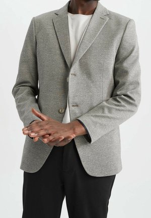Mann trägt hellgrauen Blazer über weißem Hemd und schwarzen Hosen, verschränkt die Hände vor dem Körper vor einfarbigem Hintergrund.