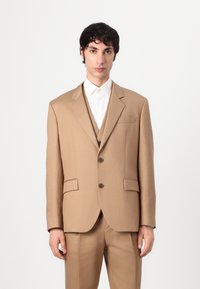 Tan suit with a notched lapel, single-breasted design, two front pockets, and brown buttons. Underneath, a white collared shirt is visible.