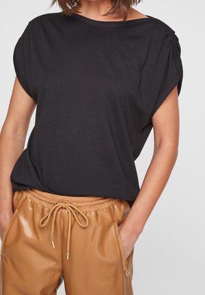 Nainen yllään väljä musta lyhythihainen paita ja vaaleanruskeat tekonahkaiset nyörillä kiristettävät housut, joissa on vetoketjulliset taskut, kädet taskuissa, rajattu hartioista.