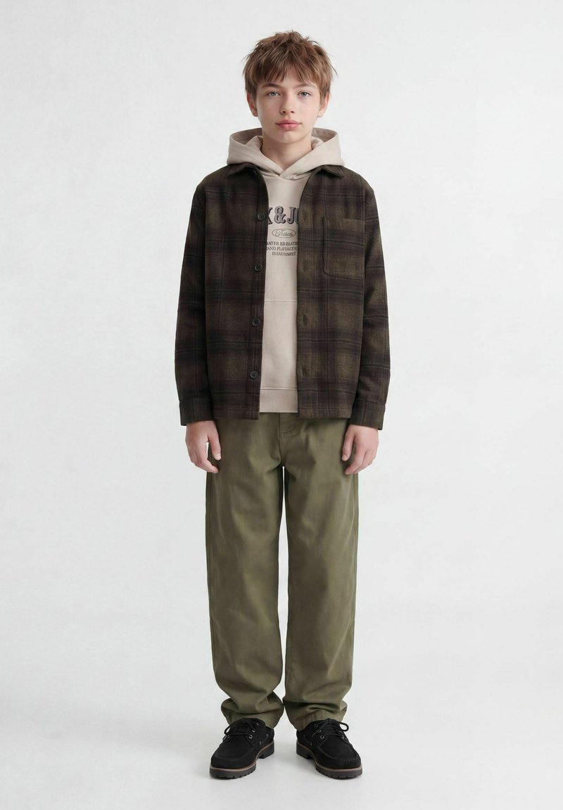 Garçon aux cheveux châtain clair portant un sweat à capuche beige sous une chemise à carreaux foncée boutonnée, un pantalon vert olive et des chaussures noires, debout devant un fond blanc.