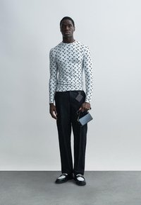 Chemise blanche à manches longues avec un motif de croissant noir, associée à un pantalon noir texturé et des chaussures noires brillantes. Tient un petit portefeuille noir.