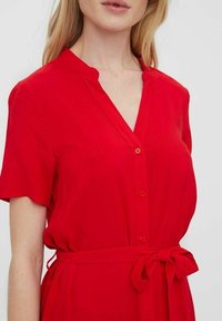 Punainen napillinen mekko lyhyillä hihoilla, v-pääntie ja sidottu vyötärö. Kangas vaikuttaa kevyeltä ja sileältä. Yksinkertainen muotoilu ilman kuvioita.