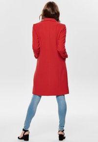 Abrigo rojo a medida con cuello estructurado, mangas largas y corte recto, combinado con jeans azul claros y tacones negros.
