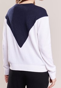 Bílý svetr s tmavě modrým trojúhelníkovým vzorem na zádech. Měkká textura, dlouhé rukávy a kulatý výstřih. Jednoduchý a ležérní styl.