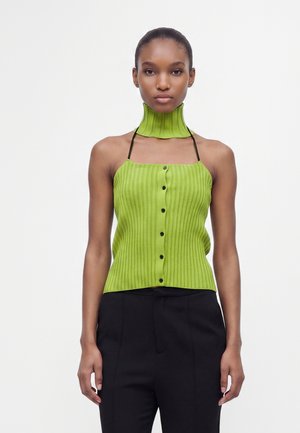 Femme portant un haut vert côtelé sans manches avec des boutons, des bretelles noires et un chauffe-cou vert côtelé assorti, associé à un pantalon noir.