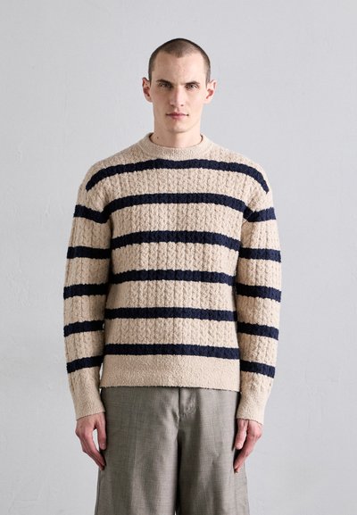 Beige pletený svetr s tmavě námořnickými vodorovnými pruhy. Texturovaná látka, kulatý výstřih a žebrované manžety. Spárováno se světle šedými kalhotami.