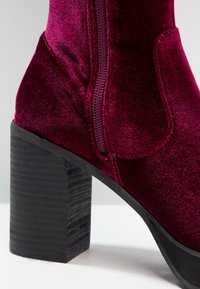 Gros plan sur une botte de cheville en velours bourgogne avec un talon noir empilé et une fermeture éclair sur le côté.