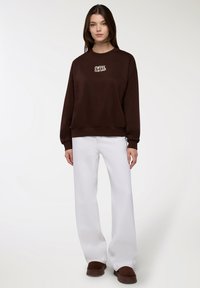 Kobieta ubrana w brązowy sweter z napisem "KOCHAM KAWOWY KLUB", białe spodnie o szerokich nogawkach i brązowe buty, stojąca na jednolitym tle.