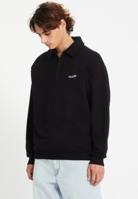 Sweatshirt zippé noir avec un col haut, comportant un petit logo blanc sur la partie gauche de la poitrine, fabriqué en tissu doux, coupe décontractée.