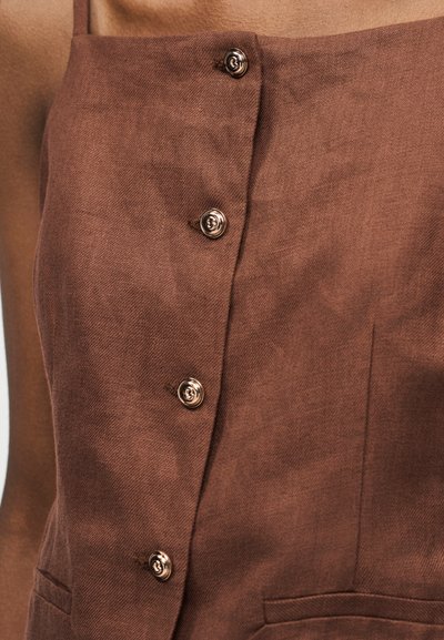 Καφέ φόρεμα χωρίς μανίκια με τετράγωνο ντεκολτέ και μεταλλικά στρογγυλά κουμπιά κατά μήκος του εμπρός μέρους, φορεμένο σε φόντο χρώματος δέρματος.