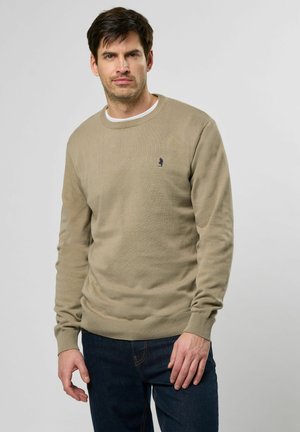Beige crew-neck sweater made of knit fabric, featuring a subtle logo in navy. Long sleeves with ribbed cuffs and hem, paired with dark jeans.