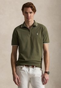 Νέος άνδρας που φοράει polo κοντομάνικο ελιάς με φερμουάρ στο γιακά, λευκό παντελόνι, καφέ ζώνη και ρολόι χειρός στο ουδέτερο φόντο.