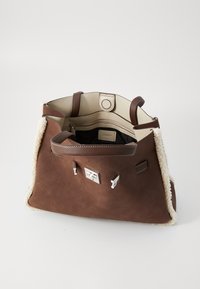 Sac fourre-tout en cuir brun avec des bordures en fausse fourrure crème, ouverture sur le dessus révélant un intérieur beige avec une poche zippée et un revêtement foncé.