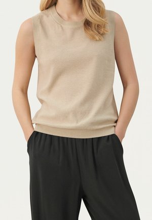 Femme portant un haut beige sans manches et un pantalon noir ample, debout les mains dans les poches devant un fond clair et uni.