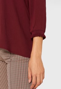 Burgundowa bluzka z rękawami o długości 3/4 i falbaniastymi mankietami, połączona z wzorzystymi spodniami w kolorze kremowym i brązowym w kratkę. Miękka, lekka tkanina.