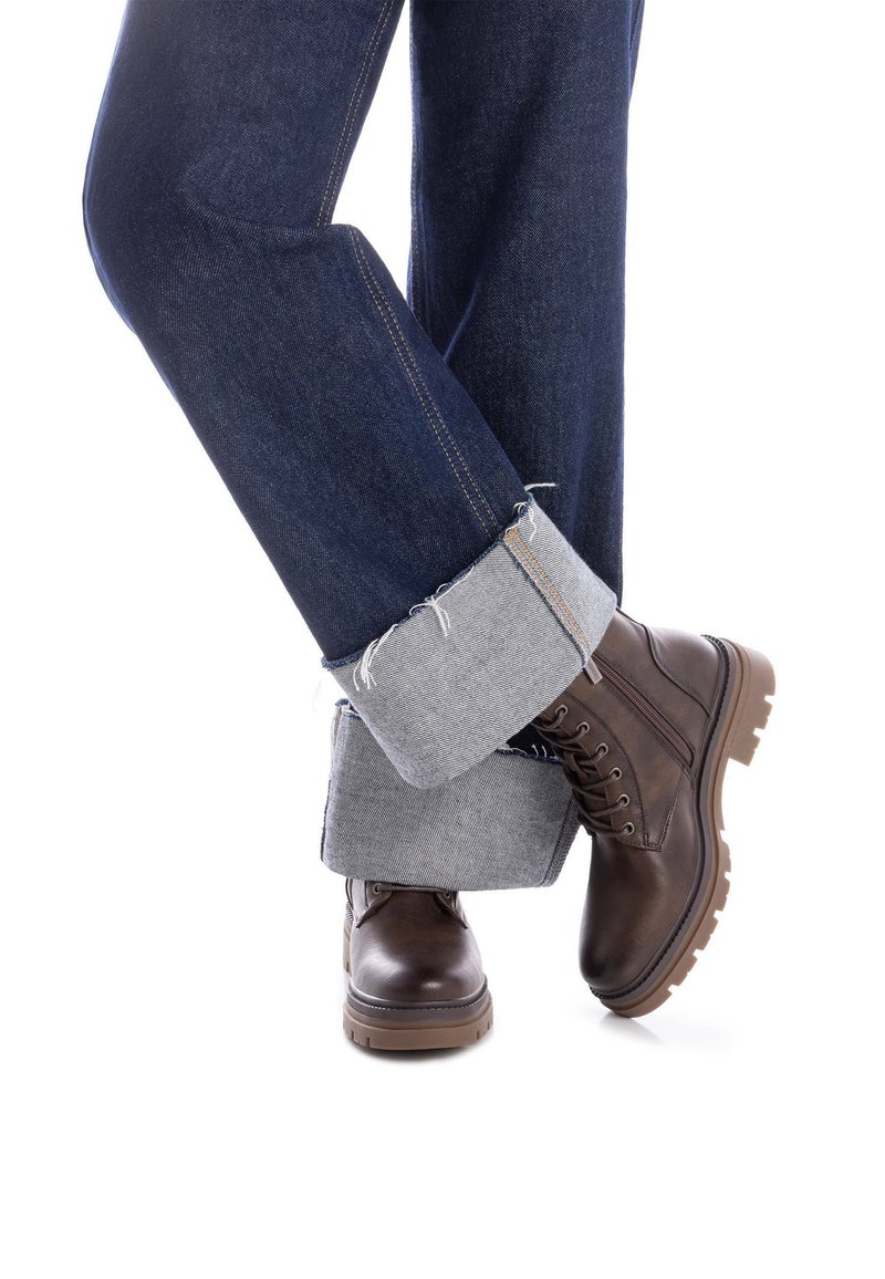 Botas de cuero marrón con suelas gruesas, combinadas con unos jeans de mezclilla remangados para exponer los puños grises. Presentan costuras en contraste y dobladillos rugosos.