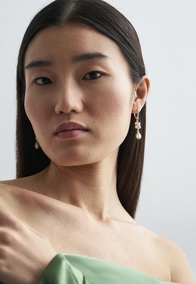 Χρυσά σκουλαρίκια με διαυγή gemstones και μια σταγόνα ως αξεσουάρ. Το μοντέλο φορεί ένα πράσινο off-shoulder τοπ μπροστά από ένα ουδέτερο φόντο.