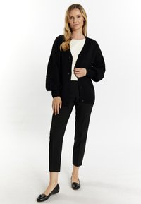 Cardigan nero a maglia con un motivo testurizzato, maniche oversize e chiusura con bottone, indossato sopra una maglietta bianca e abbinato a pantaloni neri e ballerine.