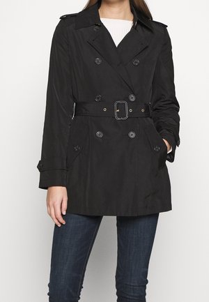 Czarny dwurzędowy trencz z paskiem, noszony na białym swetrze i ciemnoniebieskich dżinsach, z rękami w kieszeniach, stojący na jednolitym tle.