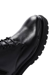 Bota de cuero negro con punta redondeada, ojales para cordones y suela de goma texturada. Presenta una superficie lisa y detalles de costura.