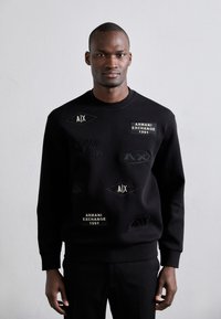 Črna dukserica z različnimi teksturiranimi patcami, ki vsebujejo logotipe "Armani Exchange" in "AIX". Okrogla ovratnica in rebrasti manšeti. Gladko blago.