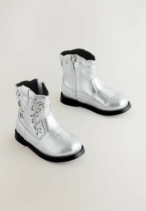 Stříbrné metalické kotníkové boty s texturovaným povrchem, dekorativními výřezy, bočním zipem a černými podrážkami.