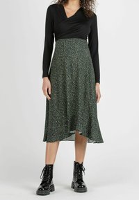 Femme portant un haut noir à manches longues, une jupe midi verte à motifs et des bottines noires brillantes à lacets, debout devant un fond uni.