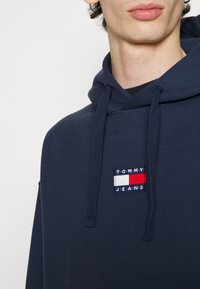 Homme portant un sweat à capuche bleu marine avec un patch logo Tommy Jeans sur la poitrine, montrant les cordons de serrage ainsi qu'une partie du cou et des lèvres.